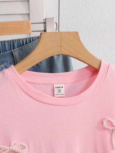 ملابس بناتي قميص T-shirt أنثوي للمراهقات من اللون الوردي بياقة مستديرة وأكمام قصيرة، بخامة مضفر بخرز ولمسة فيونكة