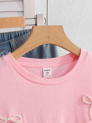ملابس بناتي قميص T-shirt أنثوي للمراهقات من اللون الوردي بياقة مستديرة وأكمام قصيرة، بخامة مضفر بخرز ولمسة فيونكة